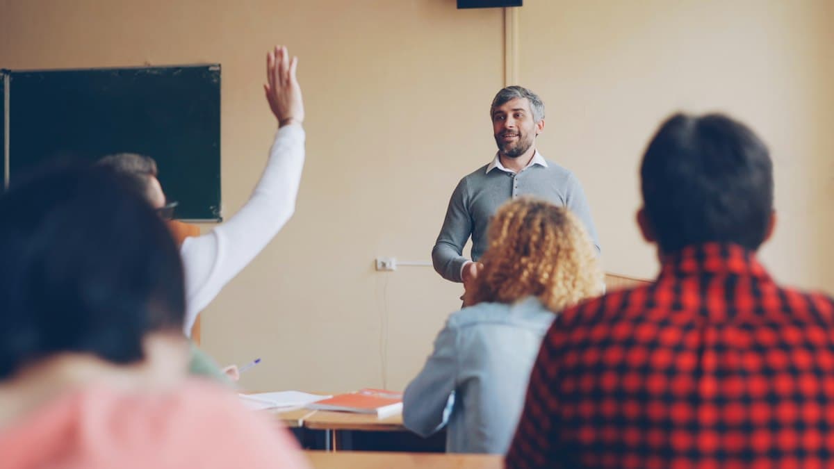 Un profesor da clase mientras una alumna levanta la mano
